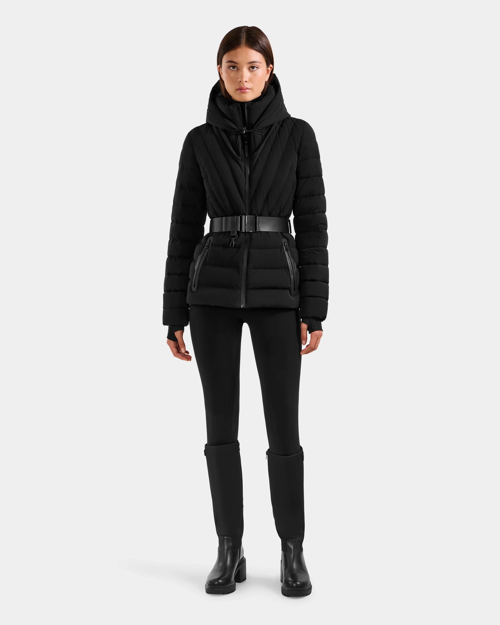A woman with straight brown hair and a neutral expression wears the Rudsak ALISA F - L125527 Black quilted puffer jacket with a belt and faux-fur trim, styled with black pants and ankle boots against a plain light gray background.