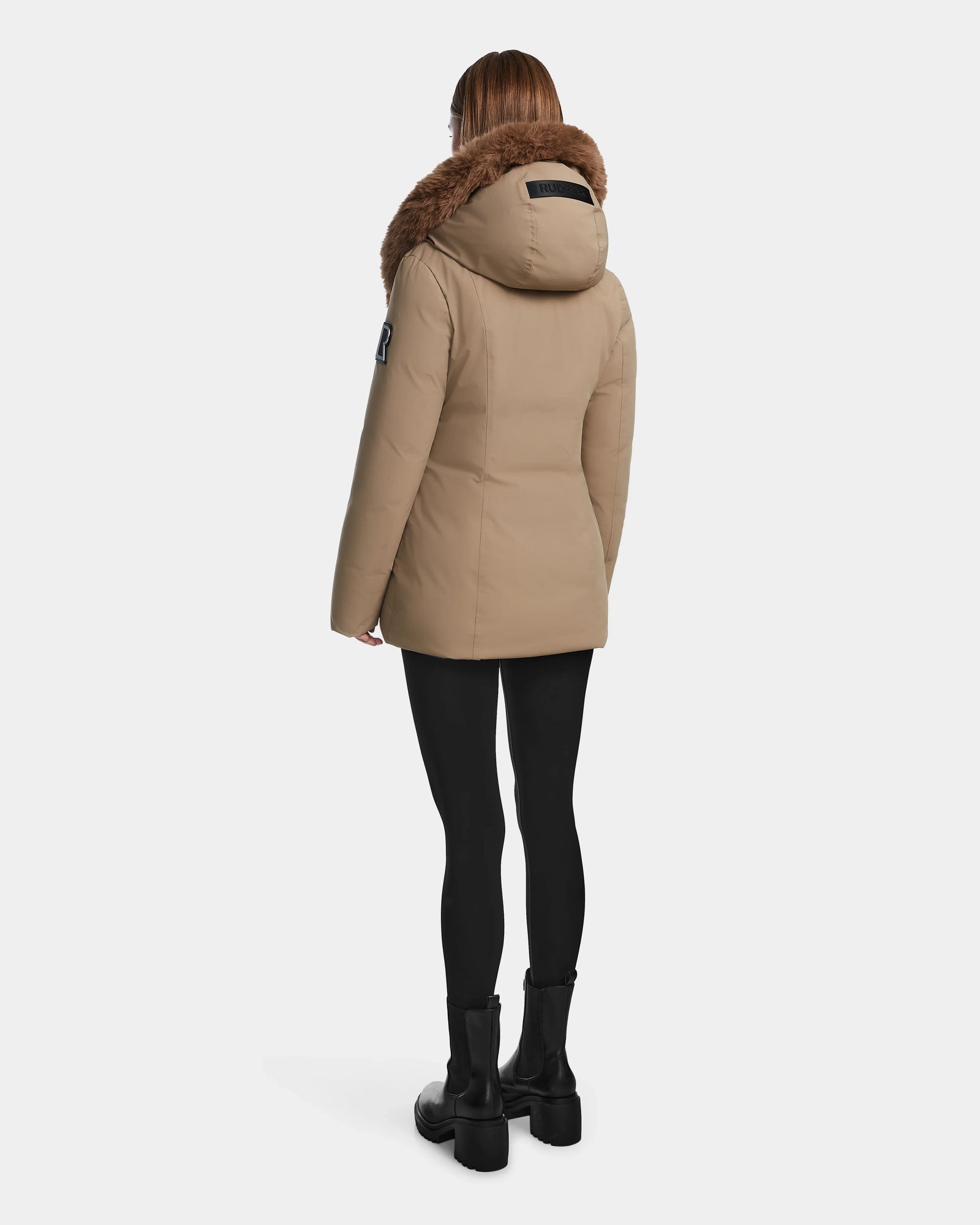 A person stands facing away against a white background, wearing the Rudsak AURORE - L123561 Sand women's beige hooded down parka with brown faux fur trim, paired with black leggings and black boots.