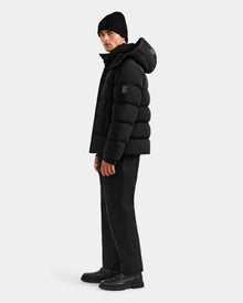 A man stands slightly sideways against a light gray background, wearing the Rudsak BRAYDEN M-S - M125524 Black men’s down puffer jacket with a hood, along with black beanie, pants, and shoes.