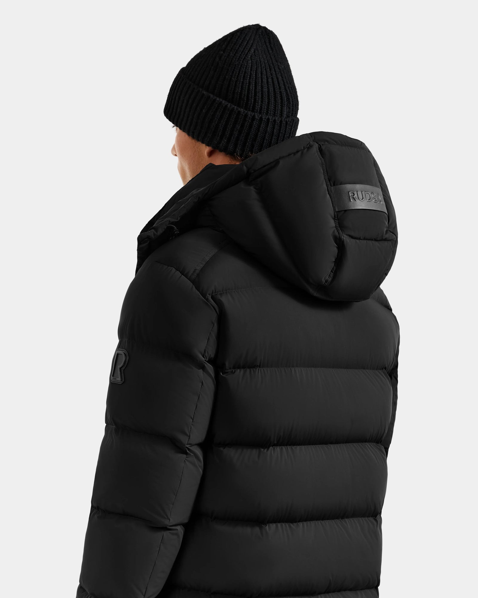 A person faces away, wearing the Rudsak BRAYDEN M-S - M125524 Black down puffer with hood and arm patch, plus a black knit beanie; "RUDSAK" branding appears on the hood.