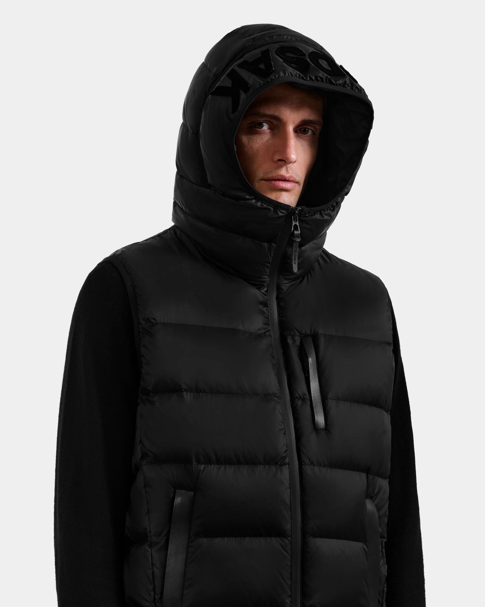 A man wears the Rudsak BROOKS - M125520 Black hooded down vest with a recycled-nylon shell over a black long-sleeve shirt, standing against a plain light background with the hood up, partially covering his head.