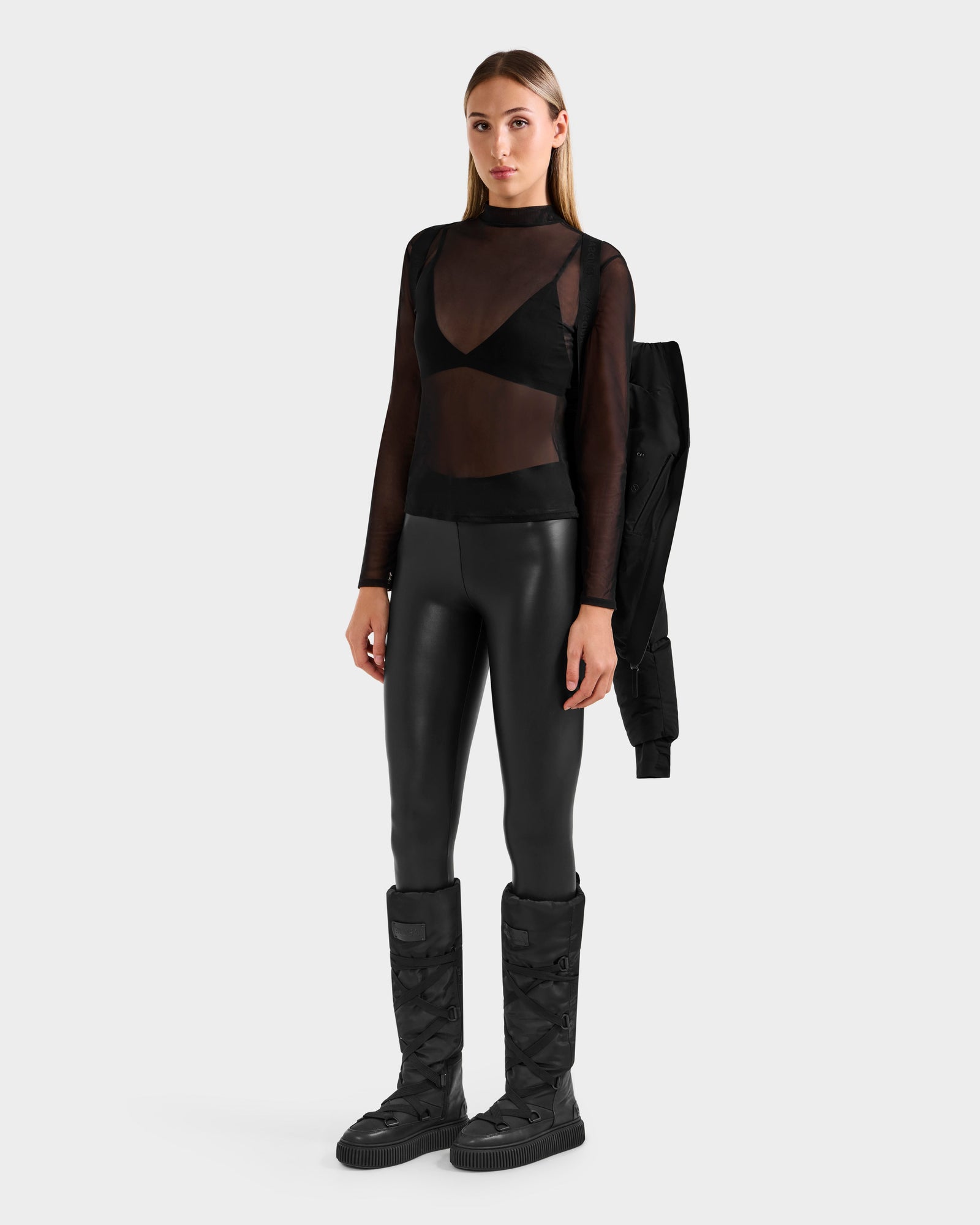 A woman in the Rudsak CHRISTELLE M - L125542 Black stands facing forward, draping a weather-resistant jacket over her left shoulder, styled with black faux leather pants and tall quilted boots against a plain light background.