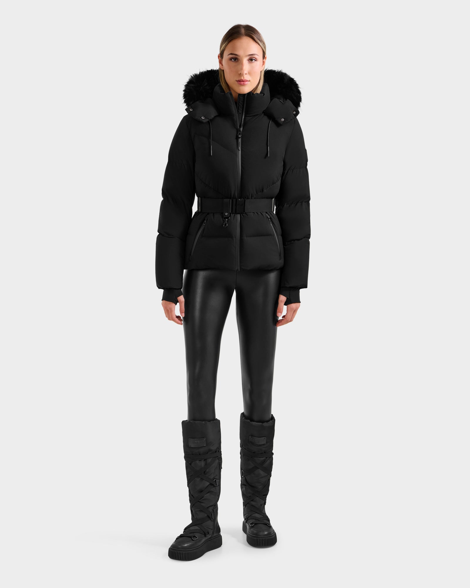 A woman faces forward wearing the Rudsak CHRISTELLE M - L125542 Black weather-resistant jacket with a fur-trimmed hood, black belted waist, leather-look leggings, and quilted boots against a plain light background.