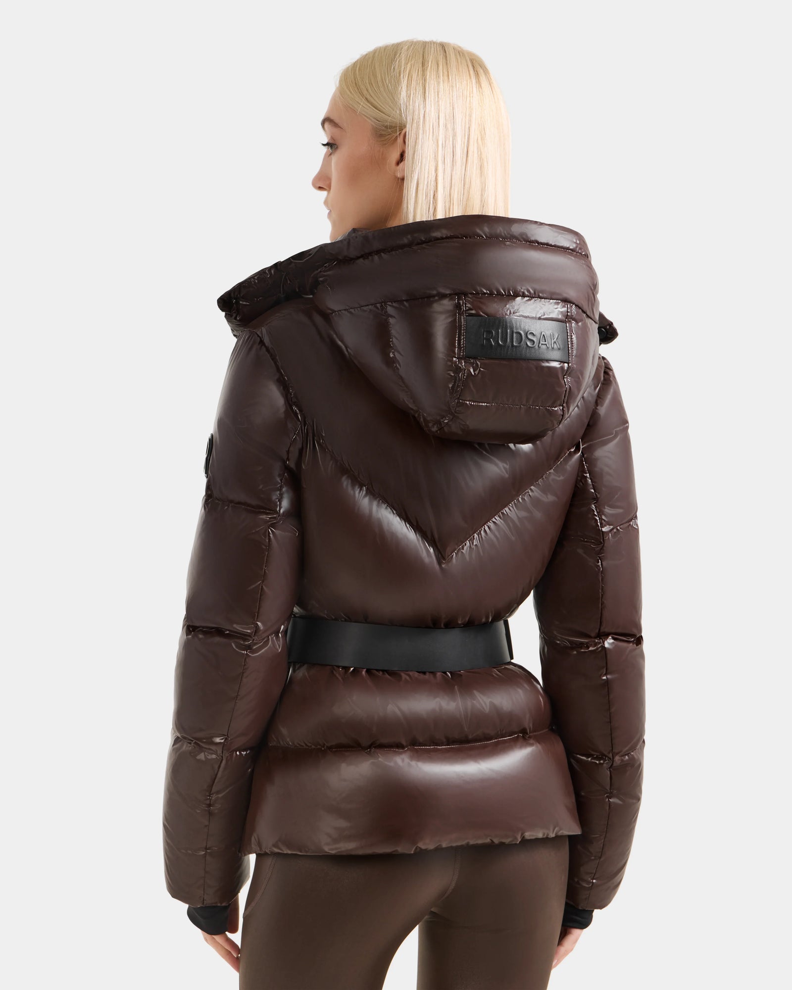 A woman with straight blonde hair is seen from the back wearing a shiny espresso CHRISTELLE S - L125544 women’s down puffer winter jacket with hood and black belt by Rudsak, featuring a rectangular brand patch on the upper back.