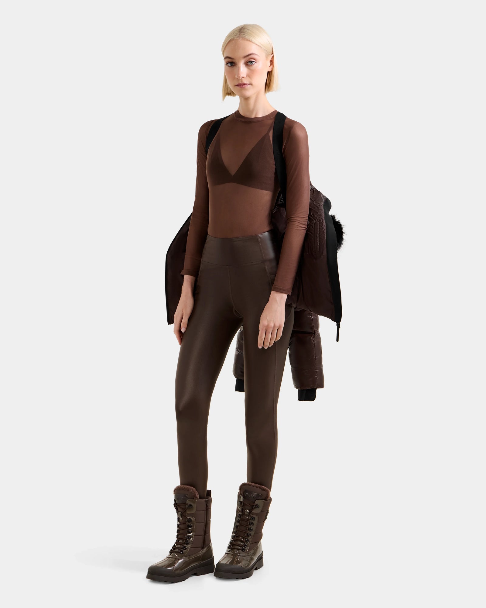 A blonde woman wears the Rudsak CHRISTELLE S - L125544 Espresso winter jacket with faux fur trim over her shoulder, paired with a sheer brown top, matching leggings, and tall lace-up boots against a plain light background.