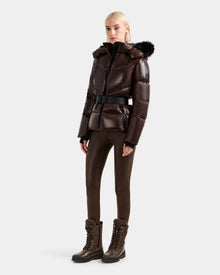 A woman wears the Rudsak CHRISTELLE S - L125544 Espresso down puffer jacket with a belt and faux fur trim hood, paired with matching brown pants and lace-up winter boots, standing against a plain light gray background.