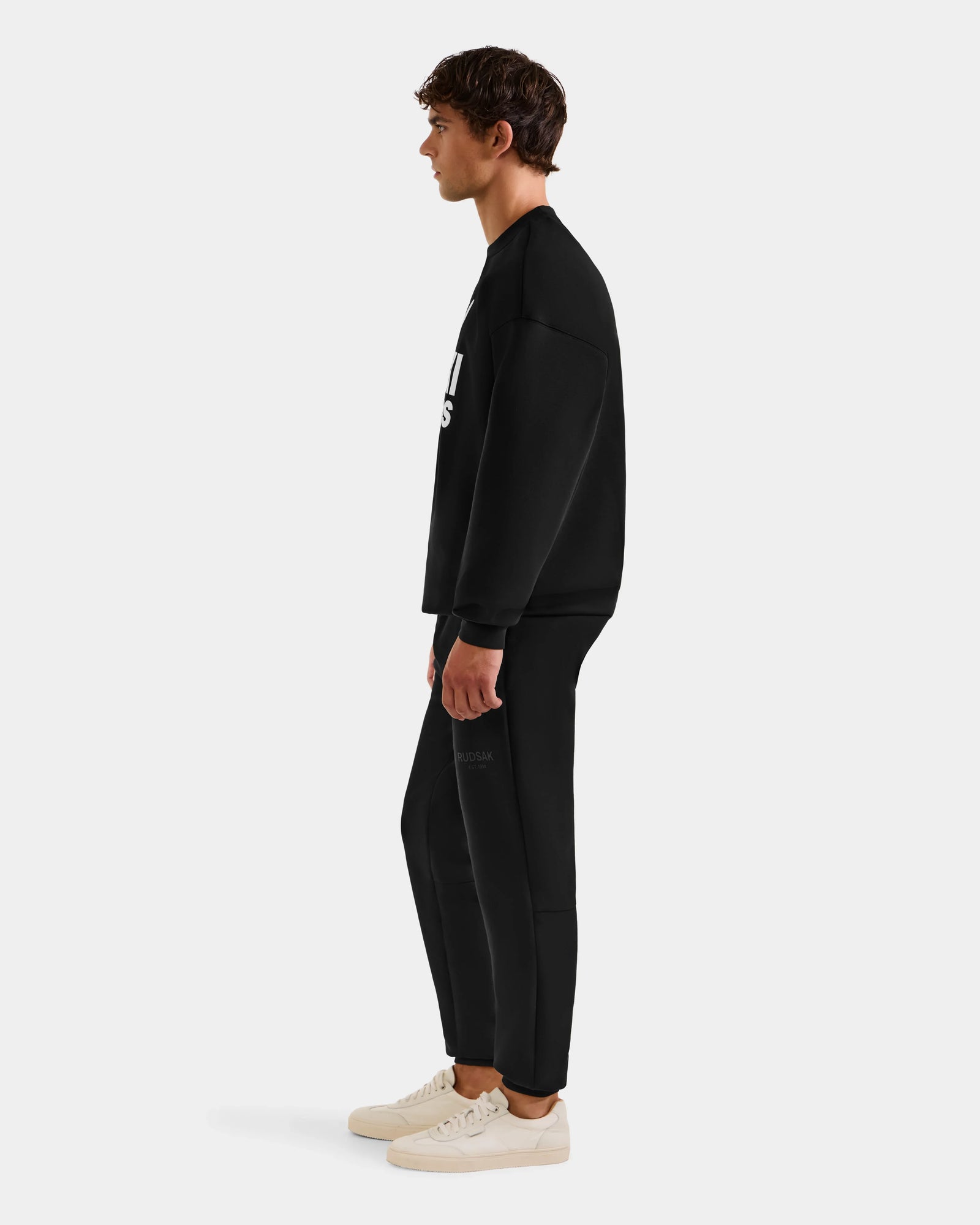 A young man stands in profile wearing Rudsak’s COLSON - M525501 Black men’s crewneck sweatshirt with matching black pants and white sneakers against a plain light gray background.