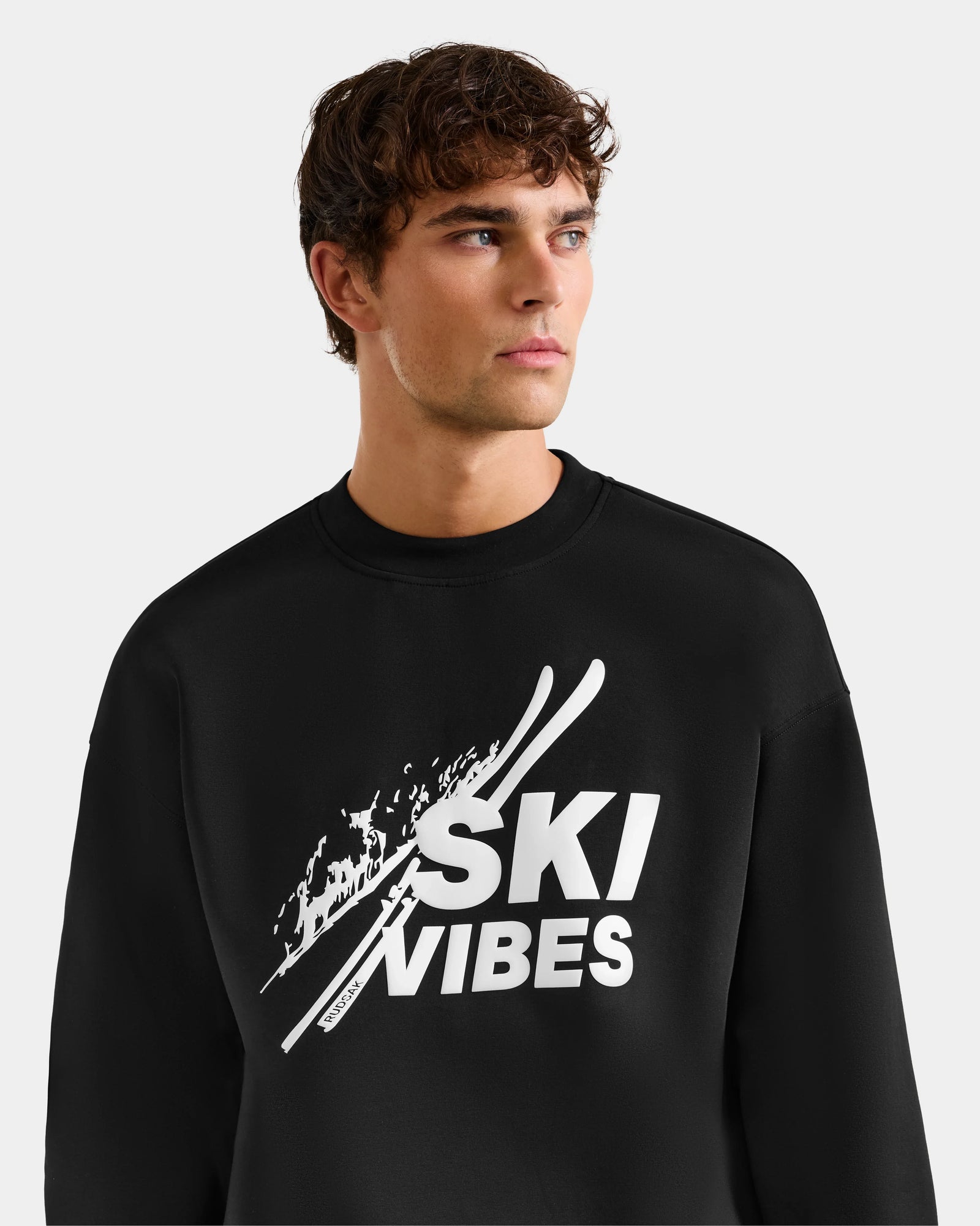 A young man with wavy brown hair wears the Rudsak COLSON - M525501 Black men’s crewneck sweatshirt, featuring the SKI VIBES logo and graphic skis, made from premium cotton-blend fleece. He looks slightly left against a plain light background.