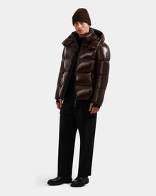 A man wearing the Rudsak DANIEL S - M124508 Espresso weather-resistant glossy shell puffer jacket, black pants, black shoes, and a brown knit beanie stands against a plain white background.