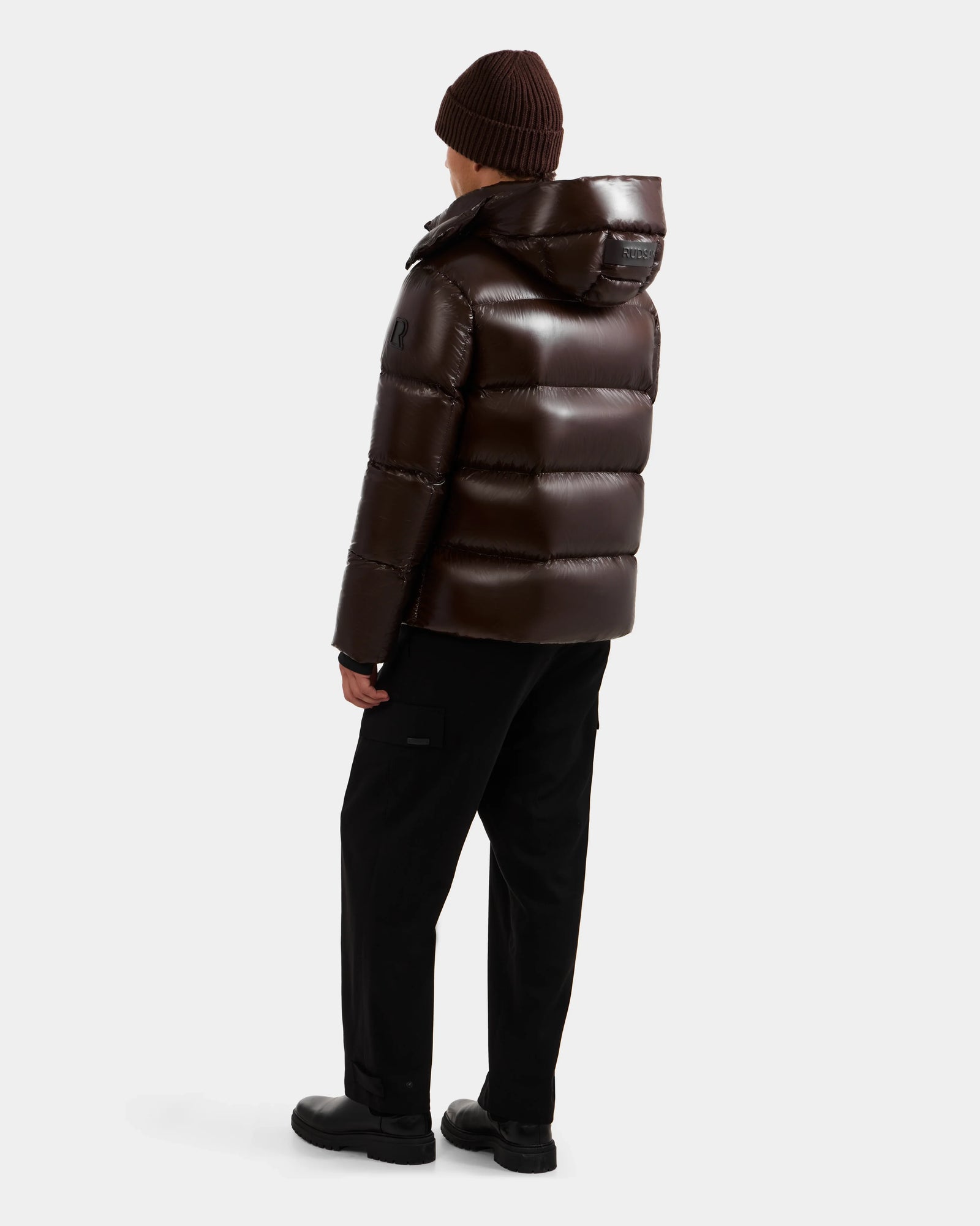 A person wearing the Rudsak DANIEL S - M124508 Espresso glossy weather-resistant puffer jacket, black pants, black shoes, and a brown knit beanie stands facing away from the camera against a plain white background.