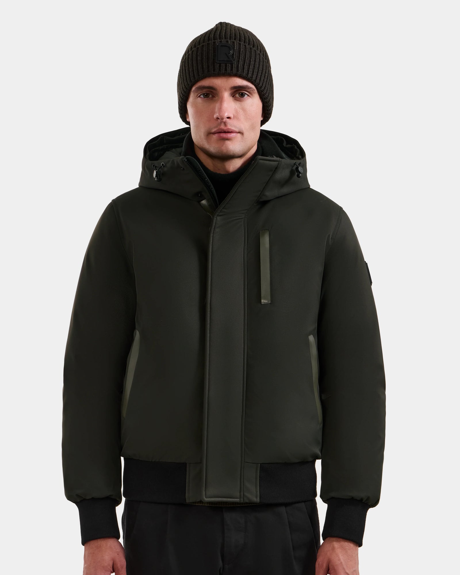 A man in a black knit beanie and the Rudsak DEACON - M123513 Dark Olive bomber-style down puffer stands facing forward against a plain light gray background.