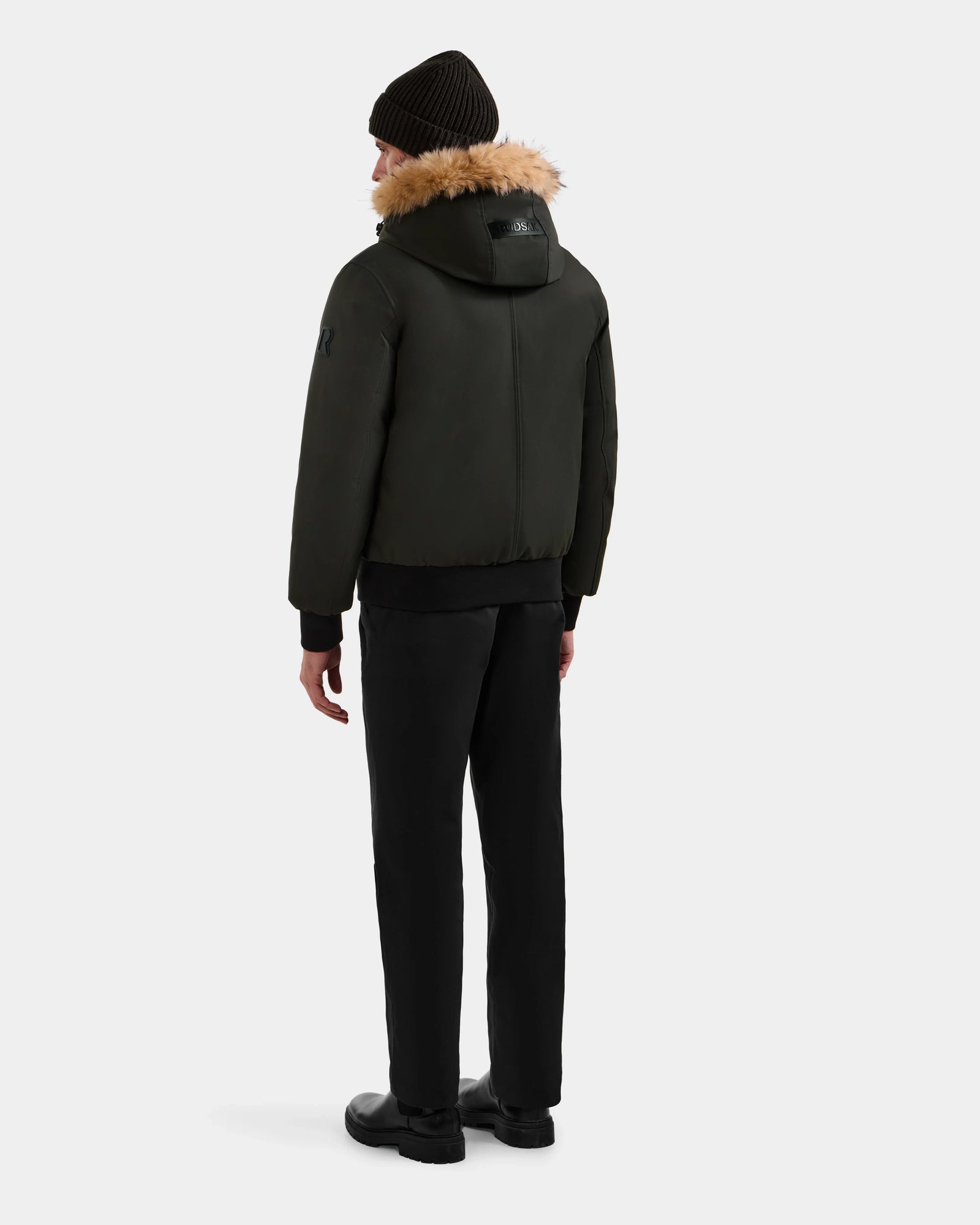 A person stands facing away against a plain light background, wearing the Rudsak DEACON - M123513 Dark Olive bomber-style down puffer, paired with black pants, black shoes, and a black knit beanie.