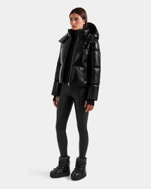 A woman wears the Rudsak DELILAH PX - L125537 Black vegan-leather puffer jacket with a hood, styled with black fitted leggings and puffer boots, standing against a plain light gray background.