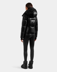 A person stands facing away, wearing the Rudsak DELILAH PX - L125537 Black vegan-leather puffer jacket, black fitted leggings, and black high-top shoes against a plain light gray background.