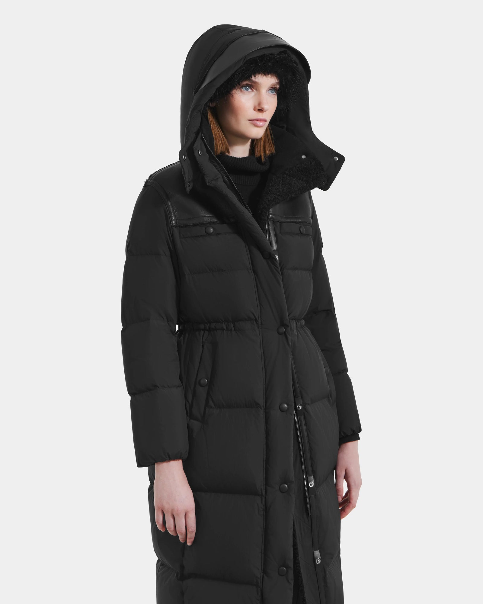 A woman stands slightly left against a white background, wearing the Rudsak EMBER M-L - L125543 Black women's long down puffer coat with a large removable hood and snap buttons.