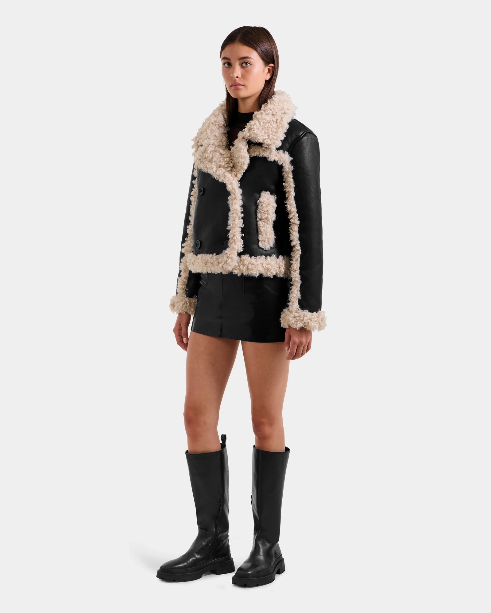 A woman with straight brown hair and a neutral expression wears the Rudsak EMERY - L125548 Black vegan-leather jacket with faux shearling trim, styled with a black mini skirt and knee-high boots, against a plain white background.