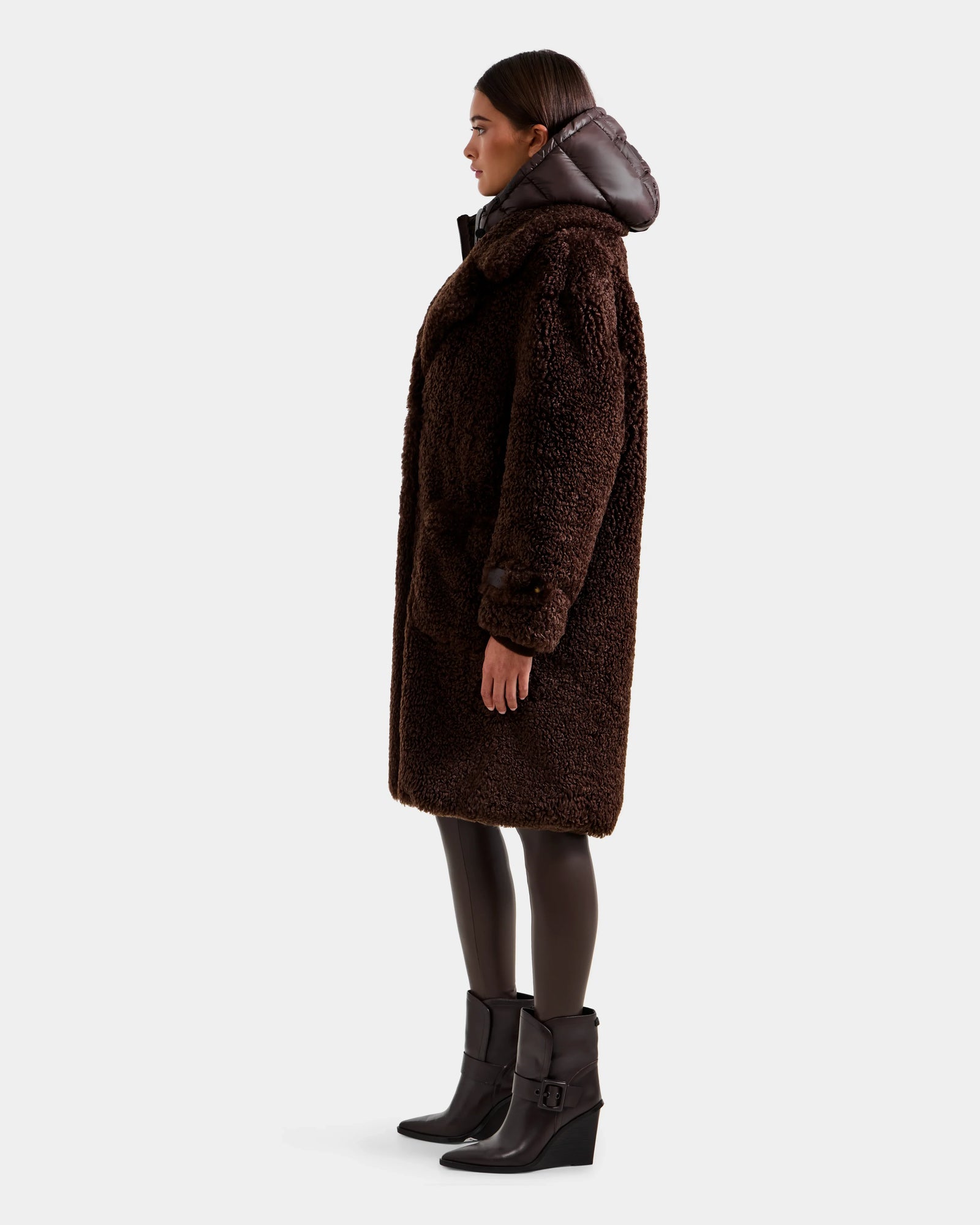 A woman stands in profile wearing the Rudsak EMRYN - L125588 Espresso, a long textured teddy jacket with a hood, paired with black leggings and wedge-heeled boots, all set against a plain light gray background.