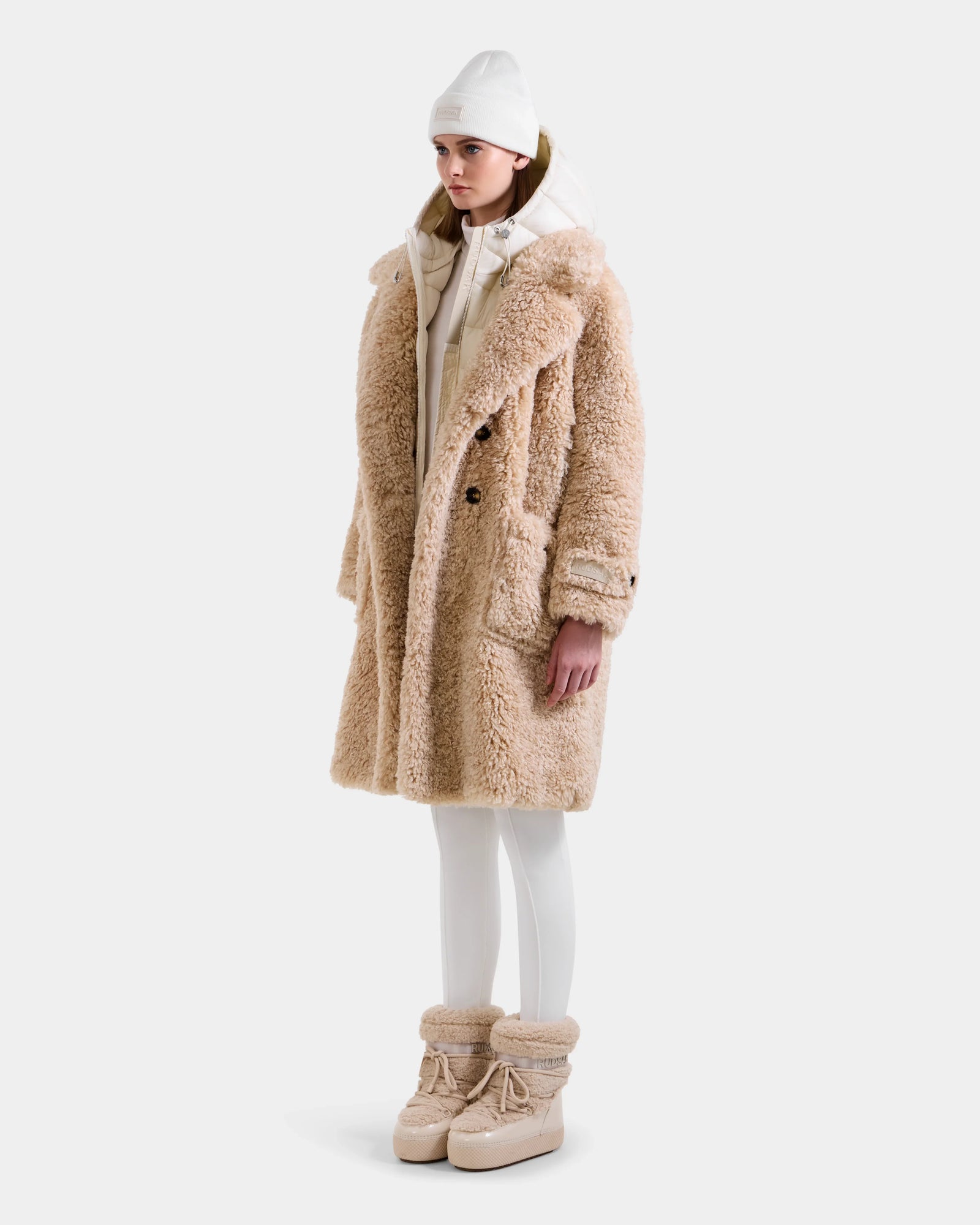 A woman wears the Rudsak EMRYN - L125588 Vanilla long beige winter coat with faux shearling, styled with white leggings, chunky beige boots, a white hoodie, and a white beanie against a plain light background.