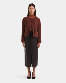A woman stands facing forward, wearing Rudsak’s FRANCESCA - 8125021 Rust cropped brown quilted puffer jacket over a brown top and a high-waisted black leather midi skirt with a front slit, paired with black pointed-toe heels.