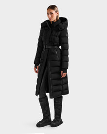 A woman in the Rudsak FRIDA M-L - L125608 Black long down puffer with detachable hood, paired with black pants and tall black quilted boots, stands against a plain light-gray background.