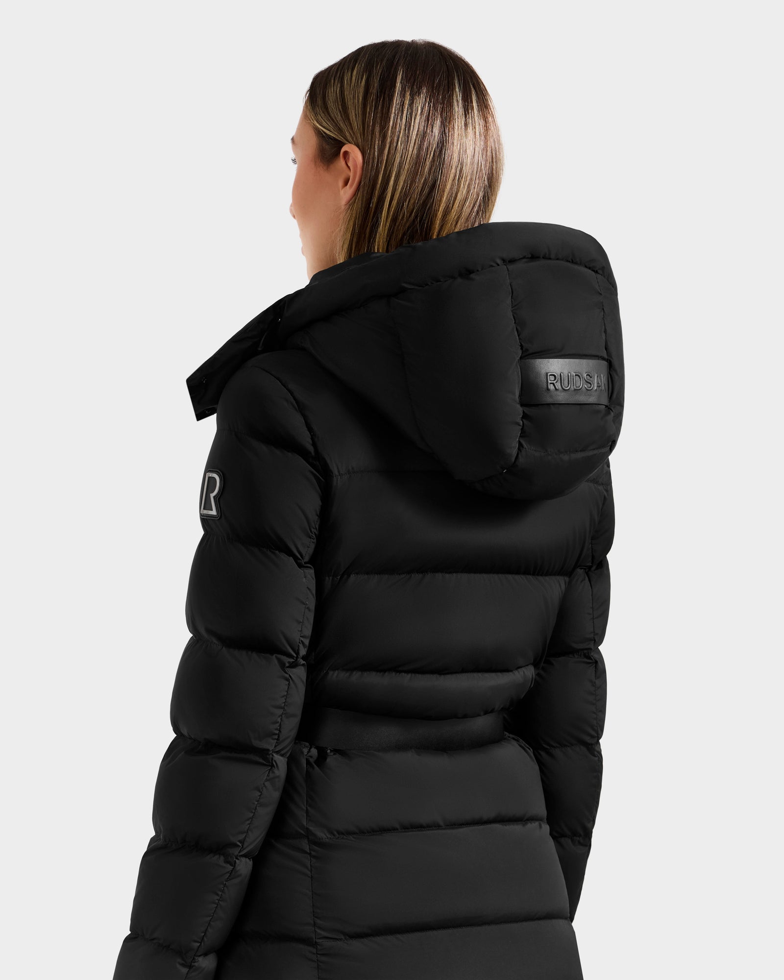 A woman with straight, shoulder-length hair models the Rudsak FRIDA M-L - L125608 Black down coat with a detachable hood, sleeve patch, and branded hood label, shown from the back against a plain light gray background.