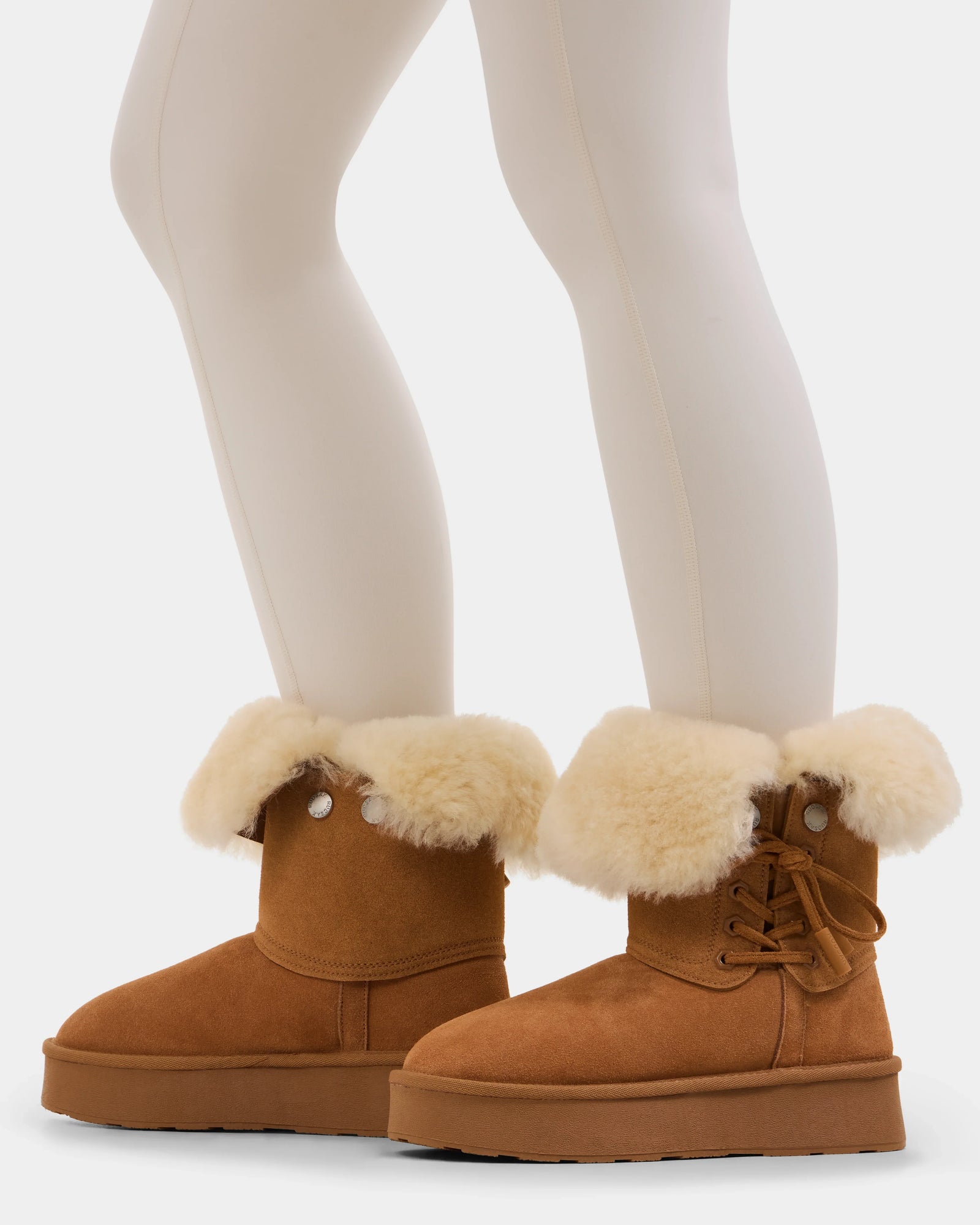 A person wears Rudsak’s INESA - L225026 Tan women’s suede platform boots with thick white shearling cuffs, side laces, and soft wool lining for extra warmth, paired with cream tights on a white background.