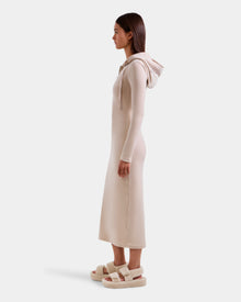 A woman in profile wears the Rudsak ISABELLA - L525502 Cream velour hooded dress with long sleeves and fuzzy sandals, featuring a subtle logo, set against a plain white background.