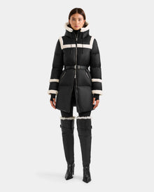 A woman faces forward in the Rudsak JULIANA L - L125535 Black/Vanilla belted puffer coat with white shearling trim, paired with black high-heeled over-the-knee boots. The background is plain white.