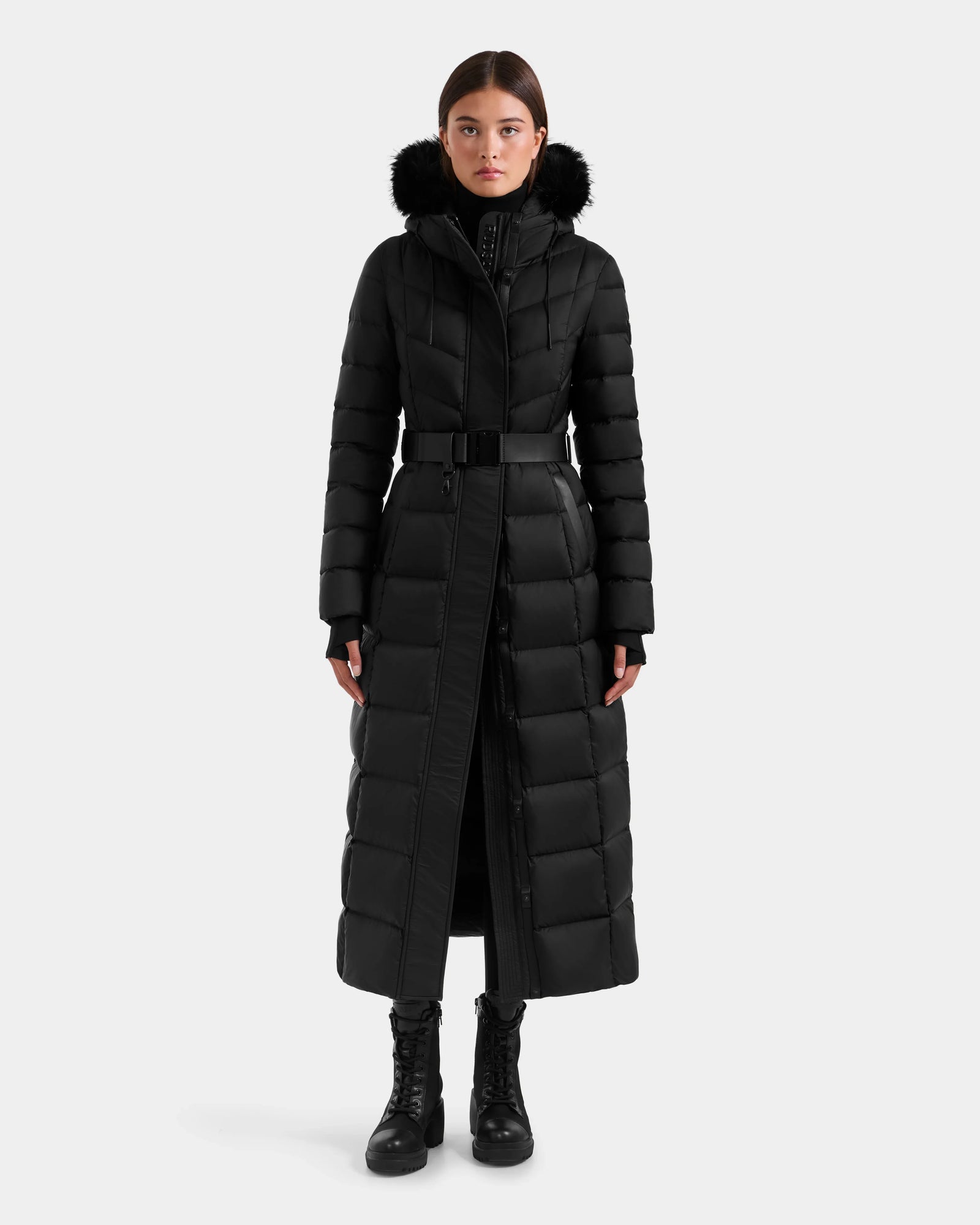 A woman faces forward against a white background, wearing the Rudsak JULIETA M-L - L125559 Black maxi down puffer with a fur-lined hood and belted waist, styled with black boots and black pants.