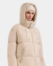 A woman wears the Rudsak KARLEE L - 8125682 Vanilla jacket, a beige puffer with vegan leather trims, drawstring hood, and zippered pockets, paired with a beige knit beanie, standing against a plain white background.