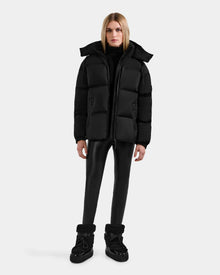 A person stands facing forward, wearing the Rudsak KARLEE M - 8125681 Black women's down puffer jacket, black turtleneck, fitted pants, and black winter boots against a plain light background.