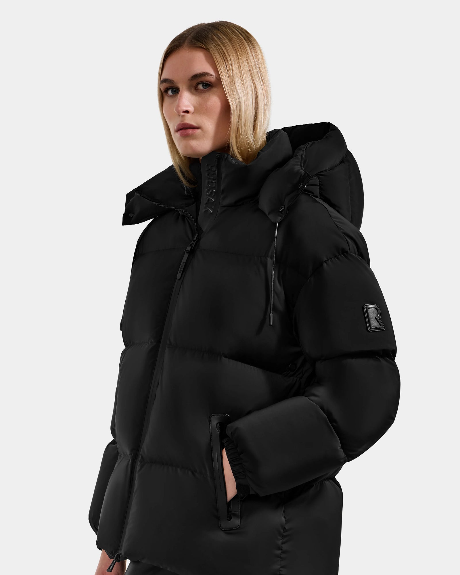 A woman with straight blonde hair wears the Rudsak KARLEE M - 8125681 Black women’s down puffer jacket with a high collar and hood, hands in pockets, standing against a light gray background—a stylish winter choice.