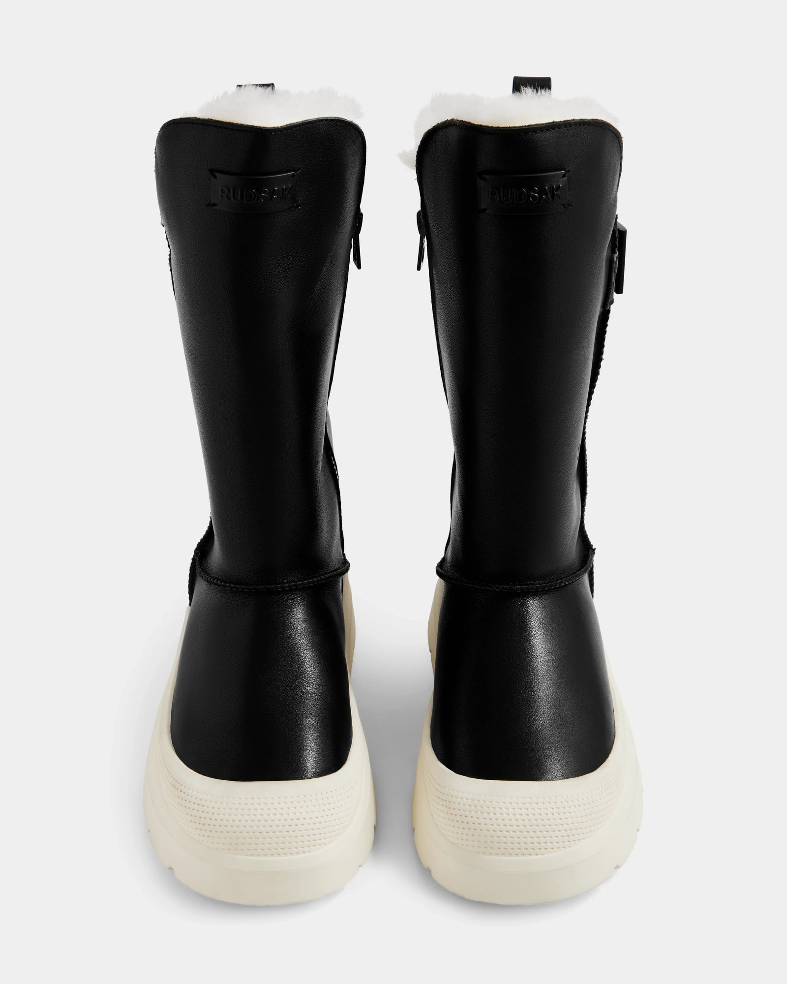 A pair of Rudsak KLAIR L - L225519 black women's leather city boots with white fur lining and thick white soles, shown from the back against a plain, light background.