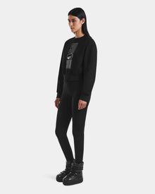 A woman faces left against a white background, wearing Rudsak's LIDDIE - L525505 Black cropped crewneck sweater with a graphic design, paired with black leggings and black lace-up boots.