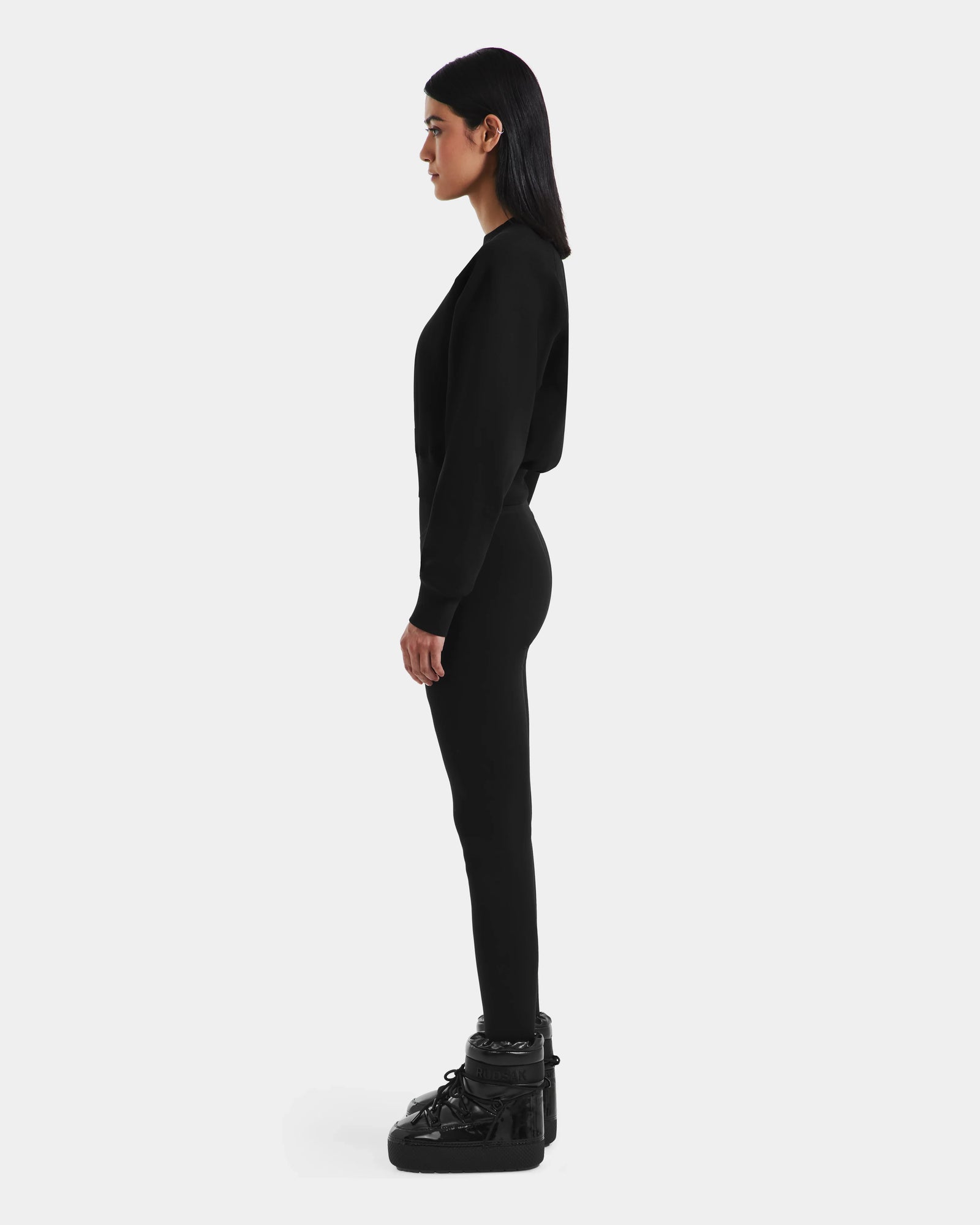 A woman with straight dark hair stands in profile, wearing Rudsak’s LIDDIE - L525505 Black fitted outfit with glossy black boots and a cropped crewneck sweater, capturing après-ski style against a plain white background.