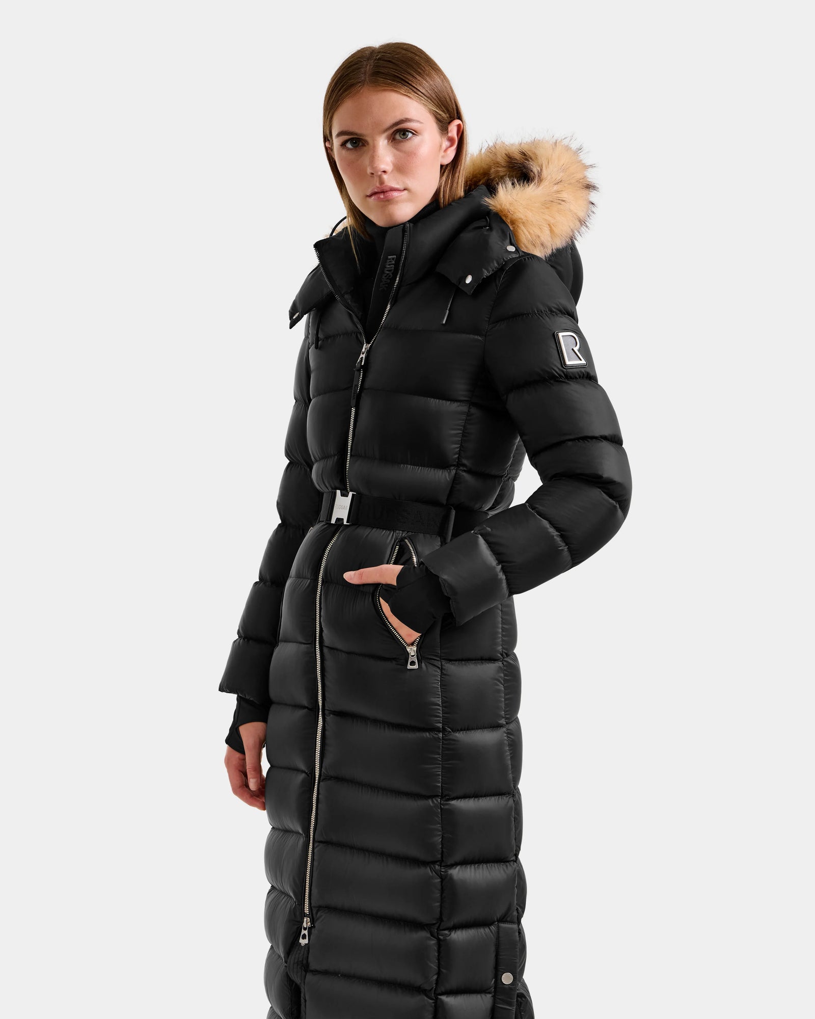 A woman wears the Rudsak LILITH L - L125528 Black long puffer coat with a tan faux fur-trimmed hood, standing against a plain light background and looking directly at the camera.