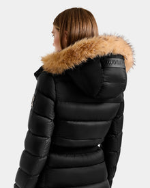 A person with light brown hair wears the Rudsak LILITH L - L125528 black puffer jacket with a fur-lined hood and horizontal stitching, featuring Rudsak branding on the hood. The background is light gray.