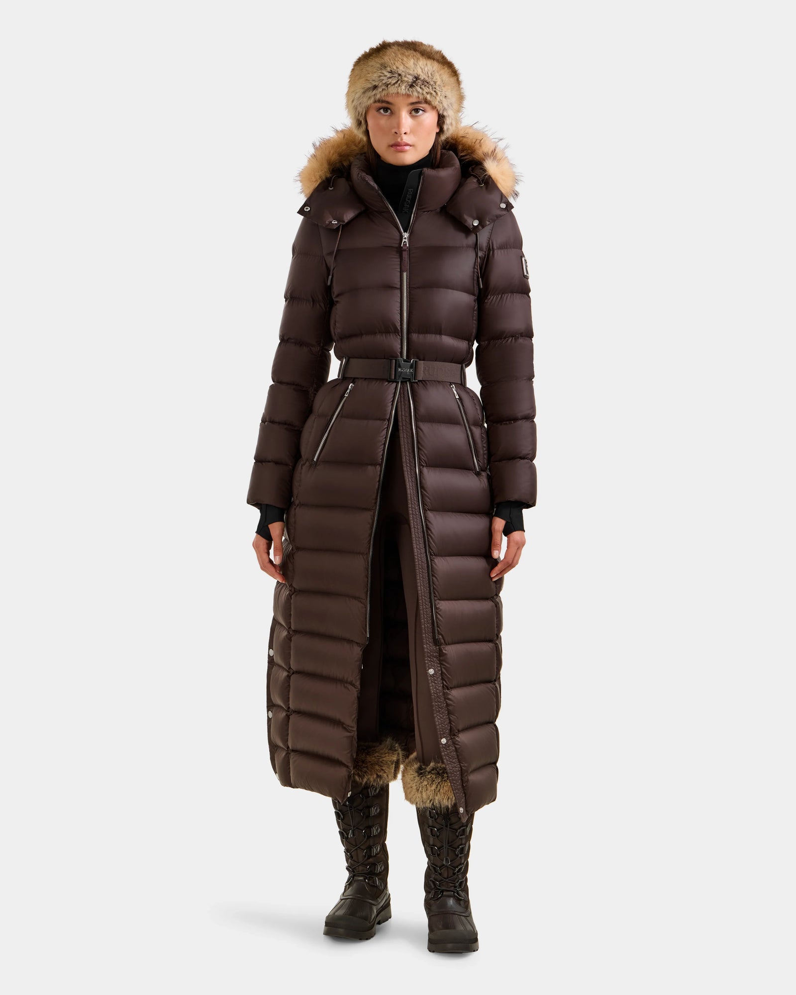 Wearing the Rudsak LILITH L - L125528 Espresso maxi down puffer with RDS-certified down, removable belt, fur-lined hood, plus a black turtleneck, gloves, winter boots, and a fur hat, a person stands against a plain light background.