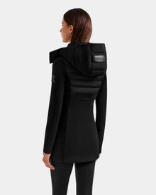 A woman with straight brown hair, wearing the Rudsak MARRY M - L125519 Black mid-length hybrid coat with a padded upper back and hood, stands facing away from the camera against a plain light gray background.