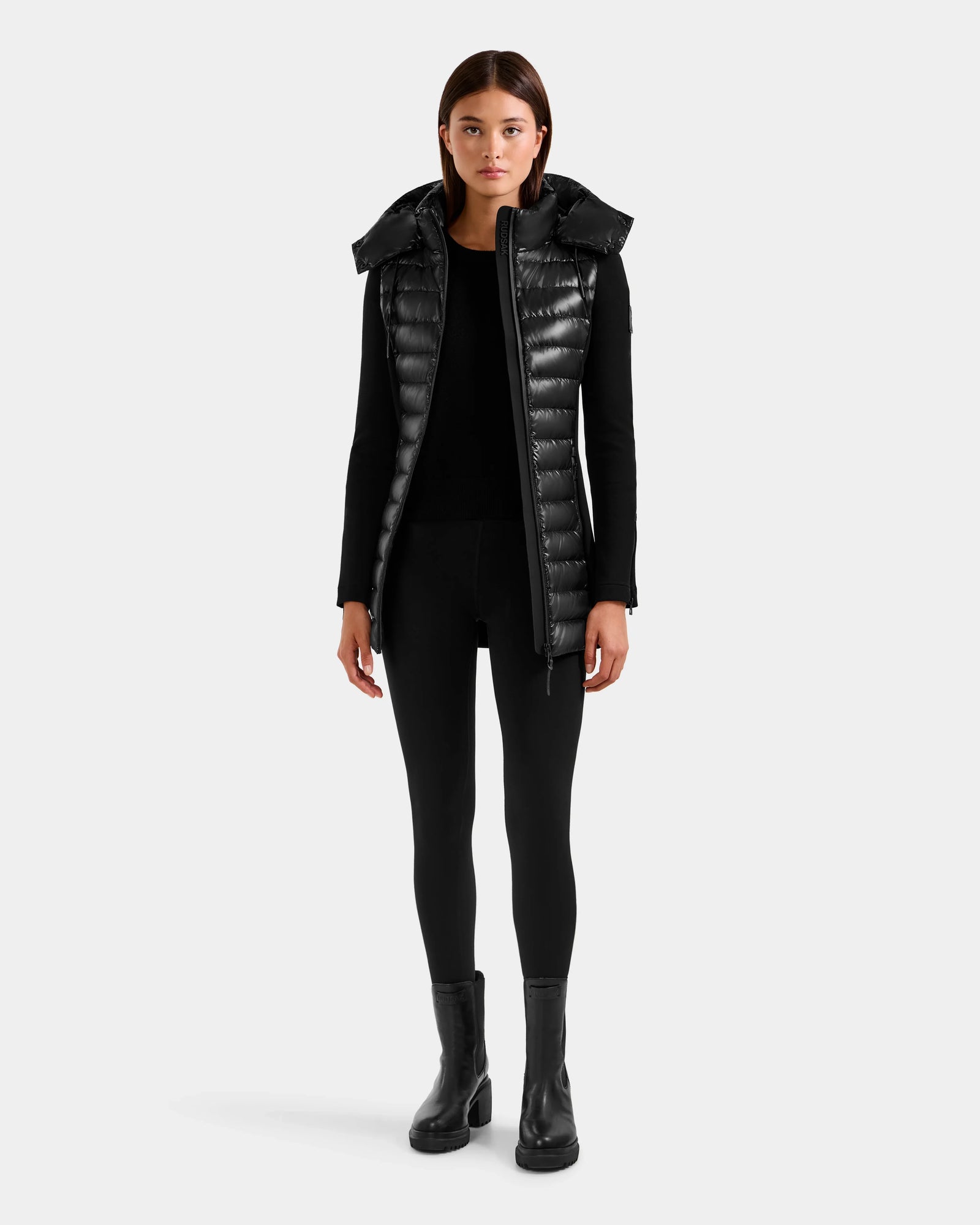 A woman faces forward against a plain light gray background, wearing the Rudsak MARRY S - L125521 Black weather-resistant long puffer vest with a high collar over black attire and heeled boots.