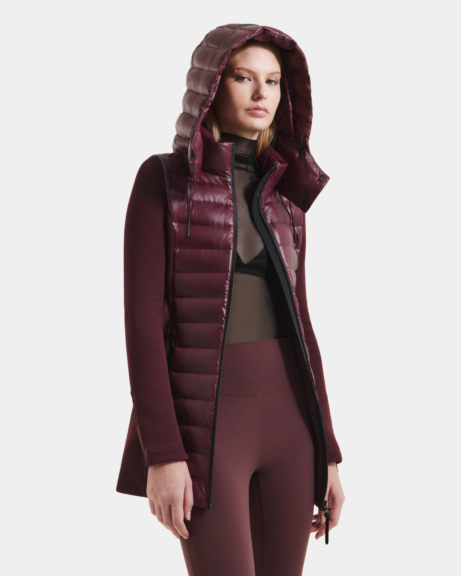 A woman wears the Rudsak MARRY S - L125521 Wine weather-resistant hooded puffer jacket with matching leggings and a sheer black top, standing against a white background.
