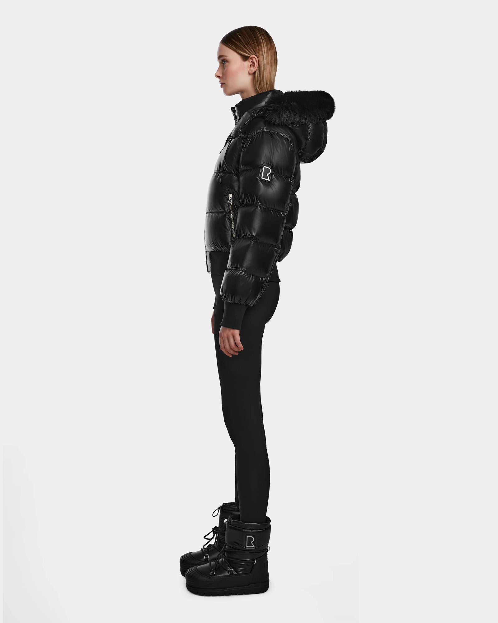 A woman in profile stands against a white background, wearing the shiny black Rudsak MAUDE Q-S - L124553 down jacket with hood, paired with black leggings and winter boots. Her straight hair is tucked behind her ears.