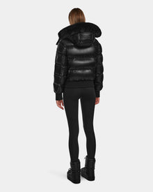 A person stands facing away, wearing the Rudsak MAUDE Q-S - L124553 Black shiny quilted down bomber with a fur-trimmed hood—this stylish winter jacket is perfect for cold days.