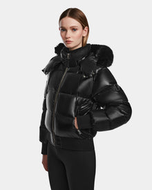 A woman wears the Rudsak MAUDE Q-S - L124553 Black, a shiny black quilted down bomber with a fur-lined hood, standing facing forward with hands by her sides against a plain light background.