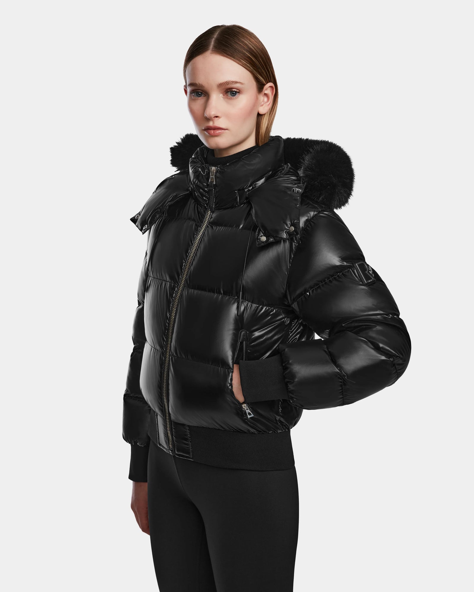 A woman wears the Rudsak MAUDE Q-S - L124553 Black, a shiny black quilted down bomber with a fur-lined hood, standing facing forward with hands by her sides against a plain light background.