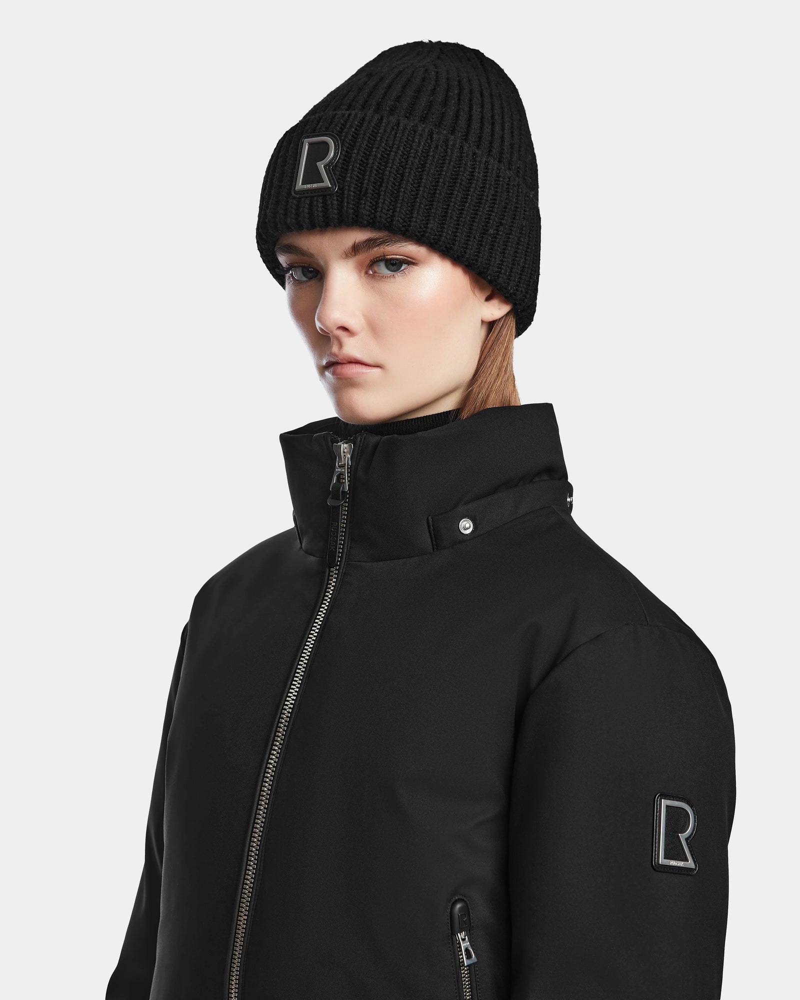 A young person wearing the Rudsak MAUDE - L124522 Black/Black winter jacket and a black beanie, both with an embroidered R logo, poses against a plain light gray background.