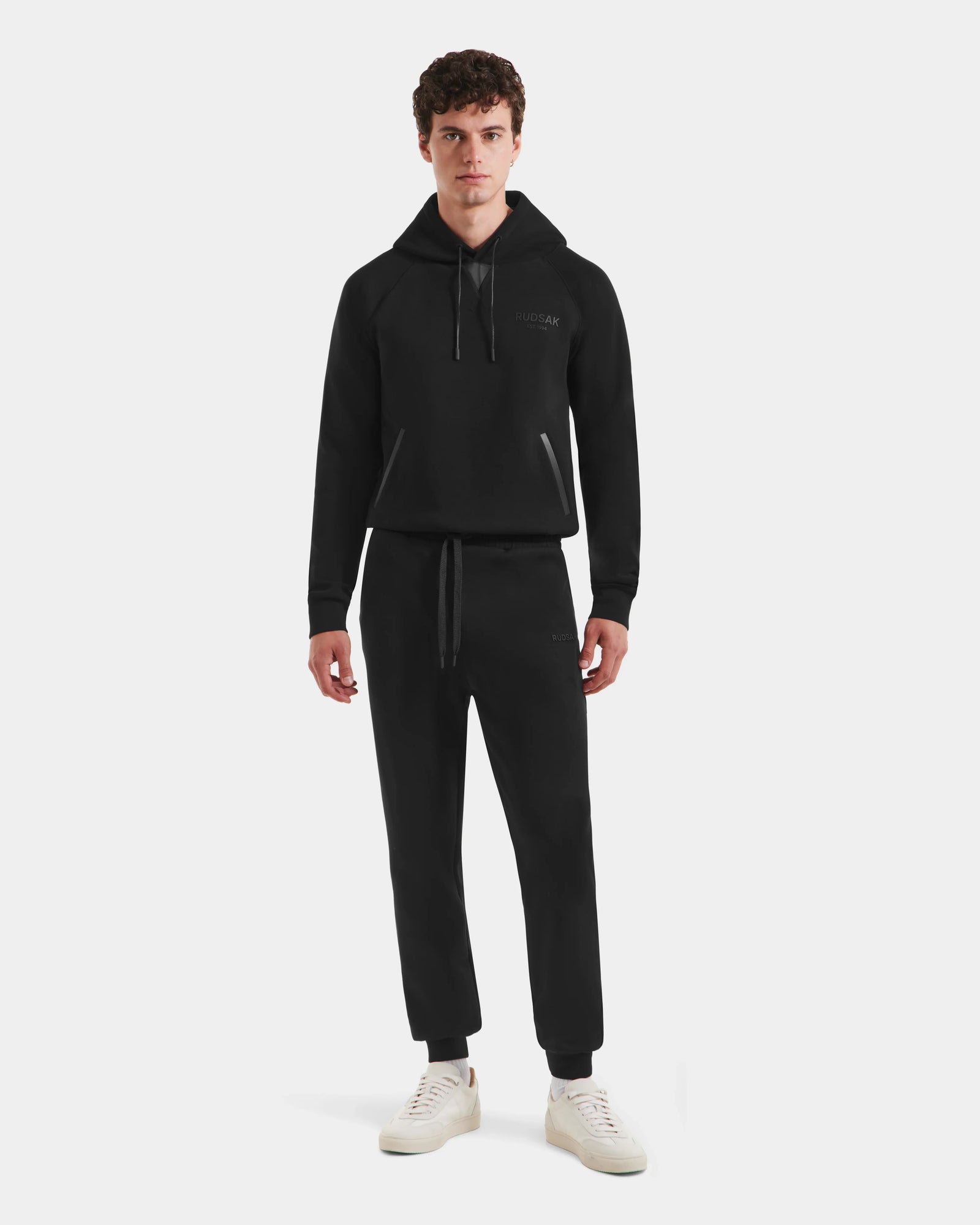 A young man faces forward in a Rudsak MAXTON - U525007 Black M unisex jogger and hoodie set with white sneakers, standing against a plain white background.