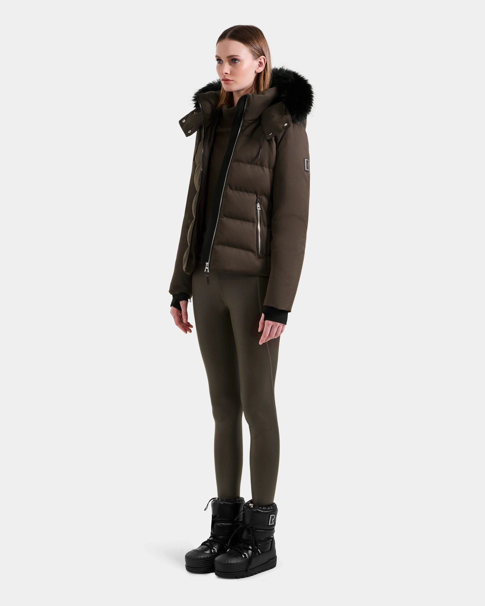A woman stands against a plain light background wearing the Rudsak MIRRY M - L124533 Espresso women's quilted down coat with a fur-trimmed hood, paired with matching brown leggings and black winter boots.