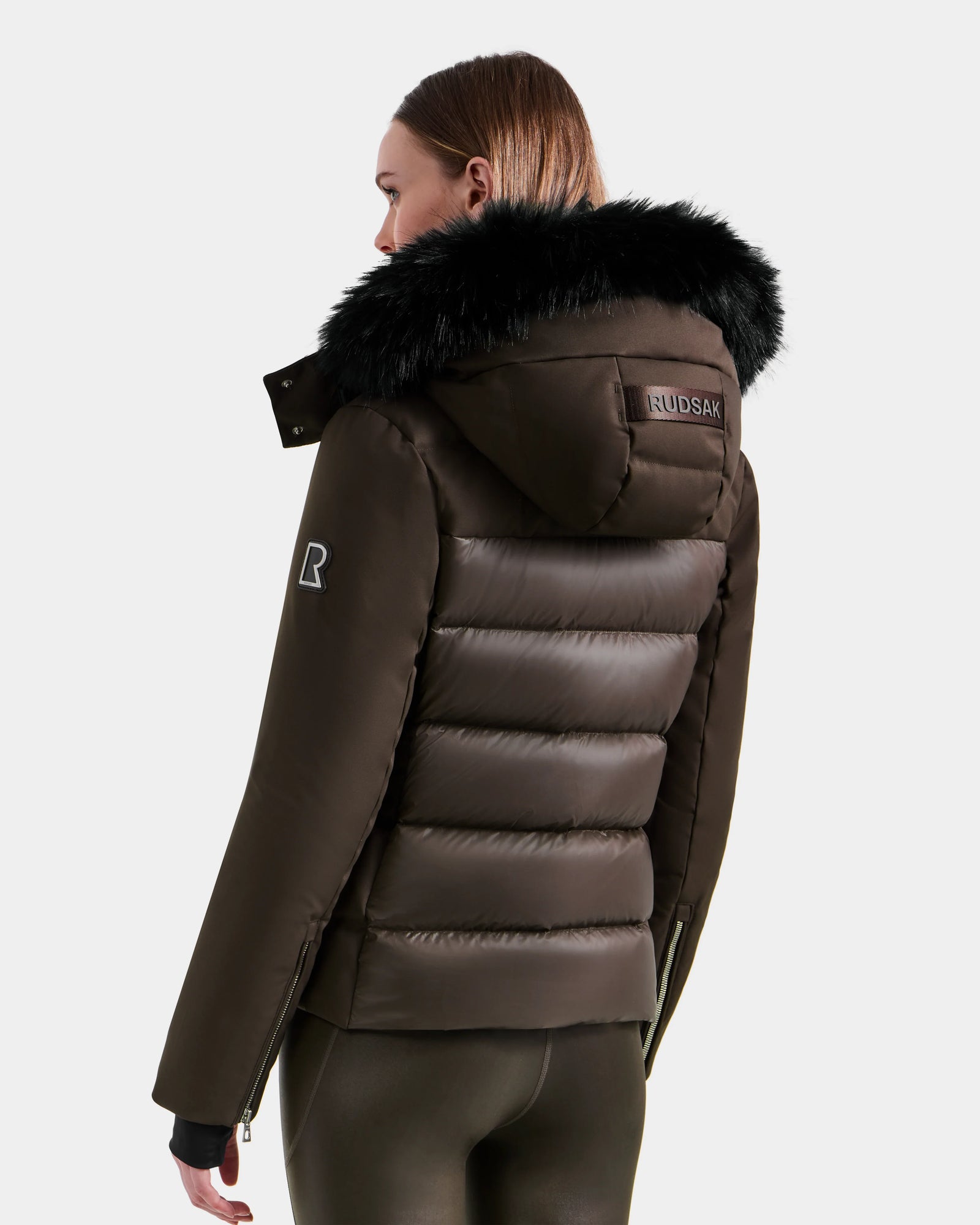 A woman is wearing the Rudsak MIRRY M - L124533 Espresso women's quilted down coat with a black fur-trimmed hood, Rudsak label on the back, and an R patch on the upper sleeve. She faces away in this stylish winter coat.