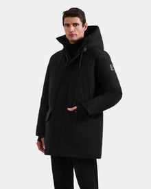 A man in the Rudsak MITCHELL - M125513 Black long men’s down parka with high collar and hood stands against a white background, one hand by his side, the other near the zipper, looking forward.